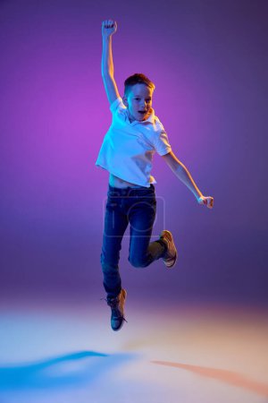 Summer break. Young boy in casual outfit jumping raising hands of joy and fun in neon light against purple gradient background. Concept of human emotions, childhood, education, fashion and style. Ad