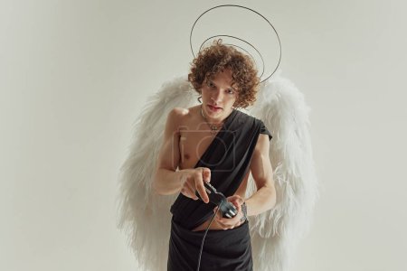 Photo for Curly-haired man with wings and halo above head looks as angel playing gamepad against white studio background. Concept of fashion and beauty, modern art and historical fiction fusion. Ad - Royalty Free Image