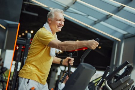 Photo for Positive, smiling mature man in vibrant sports attire with towel on shoulders work out on exercise machine at modern gym. Concept of sport, active seniors in modern life, healthy lifestyle. Ad - Royalty Free Image