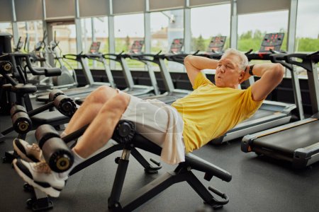 Photo for Focused on training senor man in vibrant sports gear doing abdominal exercises at modern fitness center for retirements. Concept of sport, active seniors in modern life, healthy lifestyle. Ad - Royalty Free Image