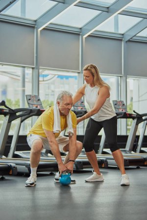 Photo for Senior man doing exercise with kettlebell and woman insures him in modern fitness center for retirements. Concept of sport, active seniors in modern life, healthy lifestyle. Ad - Royalty Free Image