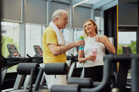 Photo for Elderly fitness duo shares conversation and hydration after workout at gym center with modern equipment. Concept of sport, active seniors in modern life, healthy lifestyle. Ad - Royalty Free Image