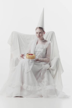 Woman in elaborate white gown and pointed hat, looks as medieval person, sits, holding sweet cake against white studio background. Concept of history, renaissance art, comparison of eras, vintage.