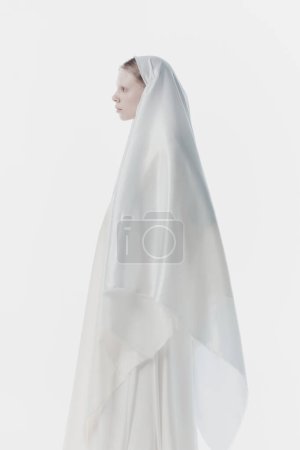 Portrait of young woman with pale skin and blonde hair, wrapped in white fabric looks as nun against white studio background. Concept of history, renaissance art, comparison of eras, vintage.