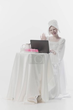 Jeune femme séduisante habillée en simple paysanne saluant tout en ayant réunion en ligne à l'ordinateur portable sur fond de studio blanc. Concept d'art de la Renaissance, comparaison des époques, vintage. Publicité