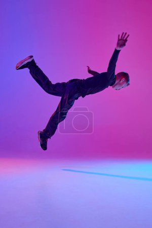 Young boy in black attire, street dancer performing tricks in mid-air in mixed neon light against vibrant gradient background. Concept of sport and hobby, music, fashion and art, movement. Ad