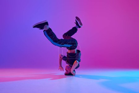 Athletic teenager, break-dancer spinning on head in motion in mixed neon light against vibrant gradient background. Concept of sport and hobby, music, fashion and art, movement. Ad