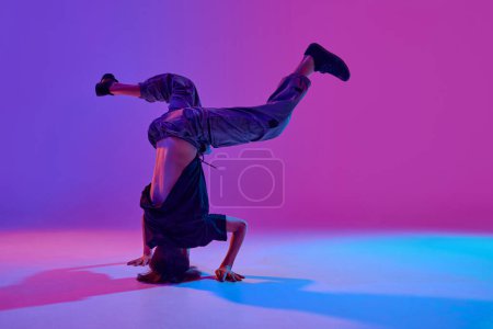 Stylish dressed young dancer, guy performing breakdance tricks in mixed neon light against vibrant gradient background. Concept of sport and hobby, music, fashion and art, movement. Ad