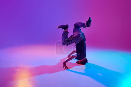 Stylish dressed young dancer, guy performing spinning on head in motion in mixed neon light against vibrant gradient background. Concept of sport and hobby, music, fashion and art, movement. Ad