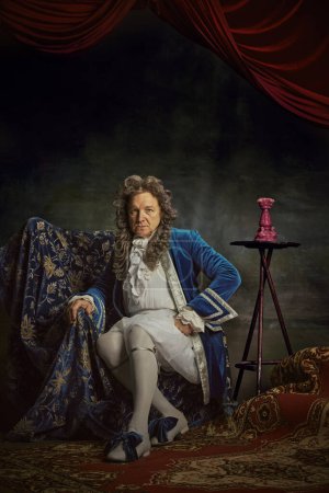 Elderly man dressed in elaborate baroque-style attire, seating regally on chair against vintage studio background. Concept of comparisons of eras, fusion of modernity and history, fashion and style.