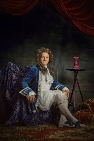 Portrait of senior man dressed in elaborate baroque-style attire, seating regally on chair against vintage studio background. Concept of comparisons of eras, fusion of modernity and history. Ad