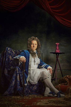 Portrait of elderly man dressed in elaborate baroque-style attire, looks as aristocratic person against vintage studio background. Concept of comparisons of eras, fusion of modernity and history. Ad