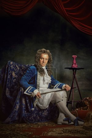 Portrait of elderly man, looks as medieval person, aristocrat, sitting on regally chair against vintage studio background. Concept of comparisons of eras, fusion of modernity and history, fashion. Ad