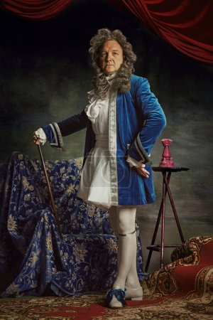 Full-length portrait of elderly man dressed as medieval person, aristocrat with confident expression against vintage studio background. Concept of comparisons of eras, fusion of modernity and history.