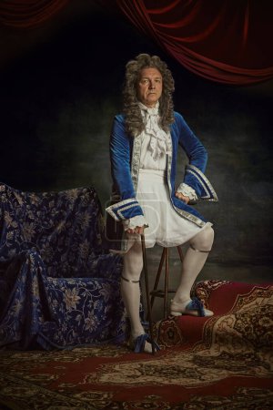 Portrait of elderly man dressed as medieval person, aristocrat with confident expression against vintage studio background. Concept of comparisons of eras, fusion of modernity and history, renaissance