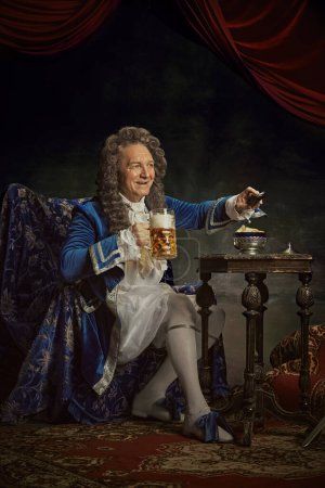 Hombre sonriente, vestidos como rey, persona medieval, tiene taza de cerveza fría y ver la televisión contra fondo de estudio vintage. Concepto de comparaciones de épocas, fusión de modernidad e historia, Oktoberfest.
