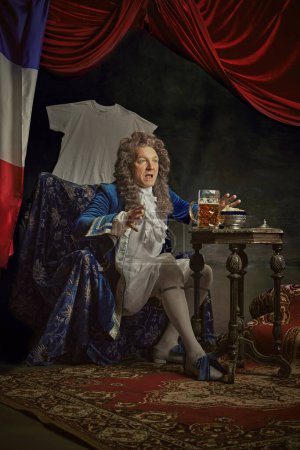 Elderly man, dressed as king in old-fashioned attire drink beer and watching sport channel on TV against vintage studio background. Concept of comparisons of eras, fusion of modernity and history. Ad