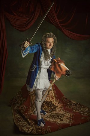 Anciano vestido con traje barroco sosteniendo espada, sentado en juguetón caballo-juguete contra fondo de estudio vintage. Concepto de comparaciones de épocas, fusión de modernidad e historia,
