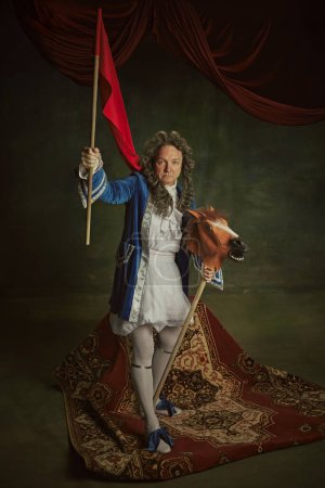 Homme âgé vêtu d'un costume baroque tenant le drapeau rouge, assis sur un cheval jouet ludique sur fond de studio vintage. Concept de comparaison des époques, fusion de la modernité et de l'histoire,