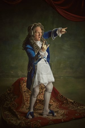 Overjoyed man dressed in richly detailed baroque costume listening music and dance in headphones against vintage studio background. Concept of fusion of modernity and history, technology.