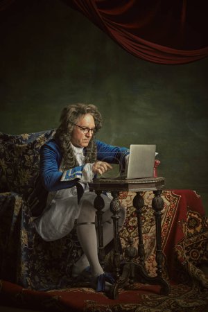 Man, dressed as king in classical attire and glasses sitting working online in modern laptop against vintage studio background. Concept of comparisons of eras, modernity and history, technology.
