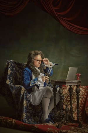 Elderly man, dressed as king in classical attire and glasses sitting watching film in modern laptop against vintage studio background. Concept of comparisons of eras, modernity and history, technology