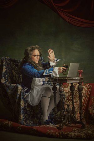King, man in classical baroque-style attire drink wine while holding meeting online against vintage studio background. Concept of comparisons of eras, fusion of modernity and history, technology. Ad