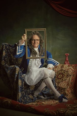 Elderly man dressed in historical baroque outfit holds up ornate picture frame against vintage studio background. Concept of comparisons of eras, fusion of modernity and history, fashion.