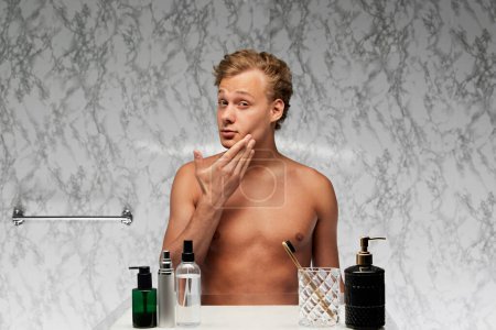 Junger Mann ohne Hemd steht im Badezimmer und blickt mit nachdenklichem Gesichtsausdruck in den Spiegel an der marmornen Wand. Konzept der männlichen Gesundheit, Schönheit und Selbstpflege, Jugend, Körper- und Hautpflege.