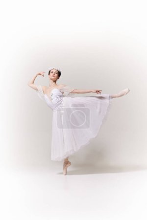 Graceful ballerina in a flowing white dress performs an elegant arabesque in pointe against white studio background. Concept of art, fusion of classic and modernity, grace and elegance.