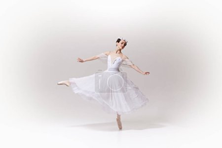 Portrait of graceful ballerina in flowing white dress, looks as fantastic character performing ballet move on pointe against white studio background. Concept of art, fusion of classic and modernity.