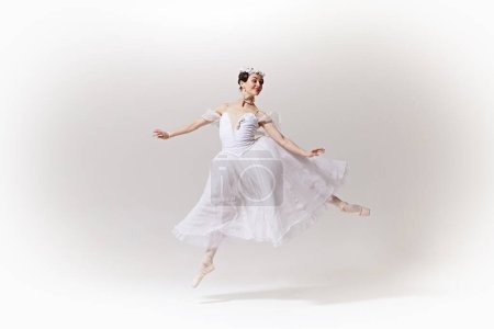 Charming ballerina in classic white dress, tutu showcasing her exquisite pose on stage against white studio background. Concept of art, fusion of classic and modernity, grace and elegance.