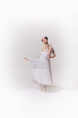 Young woman, ballerina in flowing white dress performs ballet move, conveying lightness and grace against white studio background. Concept of art, fusion of classic and modernity, grace and elegance.