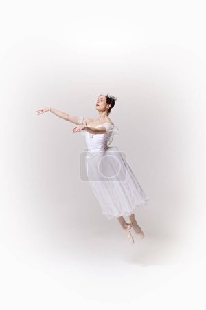 Ballerina in white flowing dress captured in mid-jump, her arms gracefully extended against white studio background. Concept of art, fusion of classic and modernity, grace and elegance.