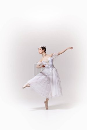 Beautiful ballerina in white dress, performing exquisite ballet move on pointe against white studio background. Concept of art, fusion of classic and modernity, grace and elegance.