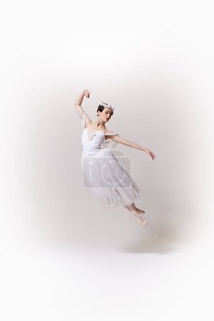 Young beautiful woman, ballerina in white costume jumps in mid-air, her movements embodying lightness and fluidity against white studio background. Concept of art, fusion of classic and modernity.