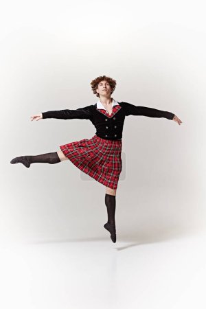 Classical ballet dancer dressed in traditional Scottish attire as fantastic character and performing against white studio background. Concept of art, fusion of classic and modernity, grace, elegance.