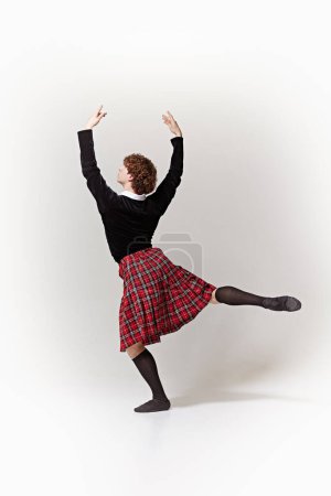 Artista de ballet clásico, con ropa tradicional escocesa, posando con los brazos levantados elegantemente sobre fondo blanco del estudio. Concepto de arte, fusión de clásico y modernidad, gracia y elegancia.
