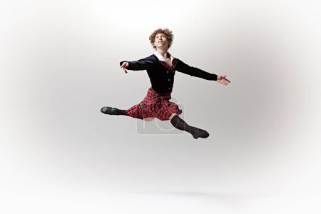Male dancer in tartan kilt and black jacket captured mid-leap, his form showcasing strength and grace against white studio background. Concept of art, fusion of classic and modernity, elegance.