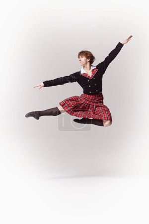 Young man, ballet dancer in traditional Scottish outfit strikes graceful pose jumping in mid-air against white studio background. Concept of art, fusion of classic and modernity, grace and elegance.