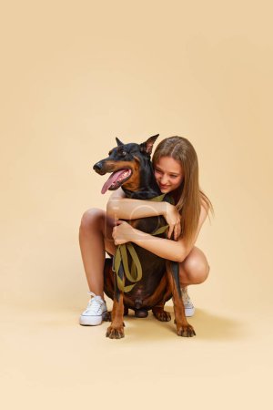 Girl with sits and hugs from behind her lovely purebred Doberman dog and smiling against beige studio background. Concept of animals and their owners, friendship, pets care. Ad