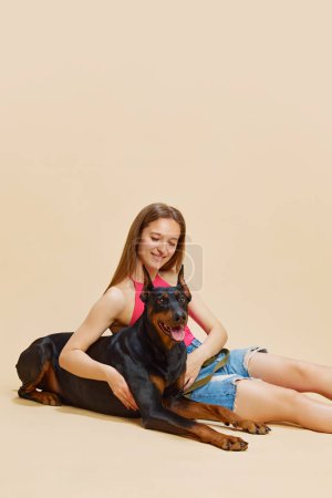 Young woman with long hair sitting on floor, smiling and hugging her large Doberman against beige studio background. Concept of animals and their owners, friendship, pets care. Ad