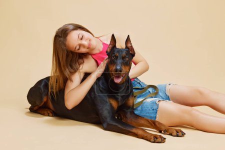 Young woman in pink top and jean shorts lies on floor cuddling with her large Doberman against beige studio background. Concept of animals and their owners, friendship, pets care. Ad
