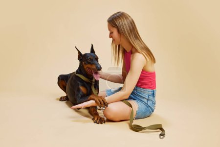 Girl in pink top and denim shorts sits on floor with her Doberman, holding pet close and smiling against beige studio background. Concept of animals and their owners, friendship, pets care. Ad