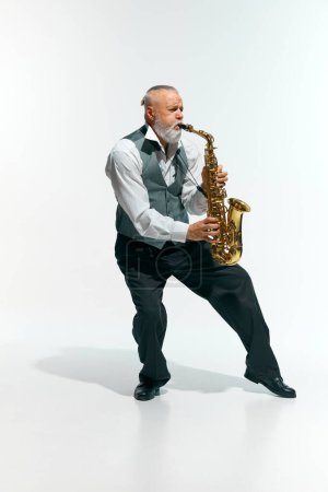 Bearded man mature man, musician informal attire playing saxophone energetically against white studio background. Concept of music, festivals, contemporary art, hobby, creativity. Ad