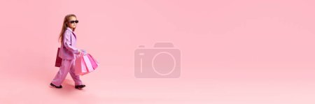 Banner. Child, girl wearing trendy clothes, carrying shopping bag against pastel pink studio background with copy space. Concept of children dreams, future career, occupation, games, fantasy. Ad