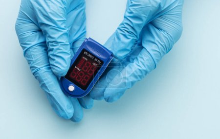 Pulse oximeter in doctor hand with glove on blue background.  A hand in a medical glove holds a device for health diagnostics