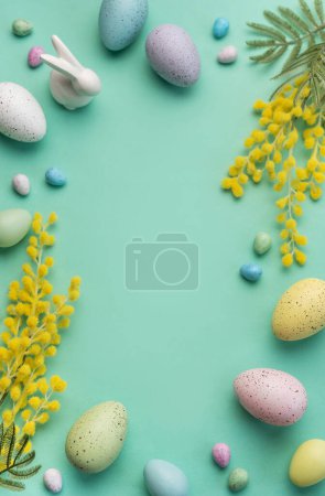 Un arrangement créatif d'?ufs de Pâques de couleur pastel, de branches de mimosa et de bonbons sur une surface verte douce, symbolisant la célébration du printemps.
