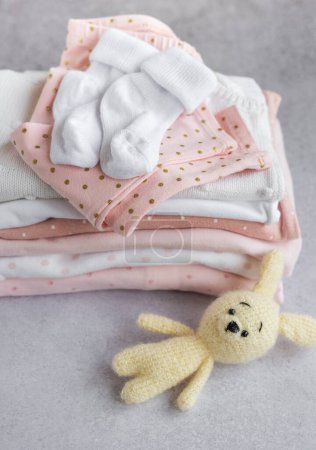 Foto de Pila de trajes de bebé sobre un fondo gris. Bebé de ganchillo juguete de conejito al lado de una pila de mamelucos para niños - Imagen libre de derechos