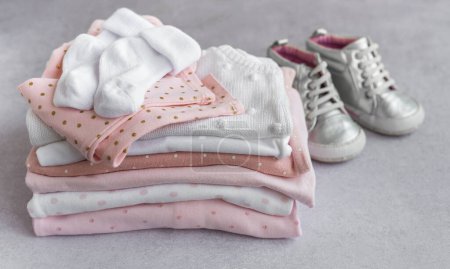 Foto de Pila de trajes de bebé sobre un fondo gris. Zapatos de bebé al lado de una pila de mamelucos para niños - Imagen libre de derechos